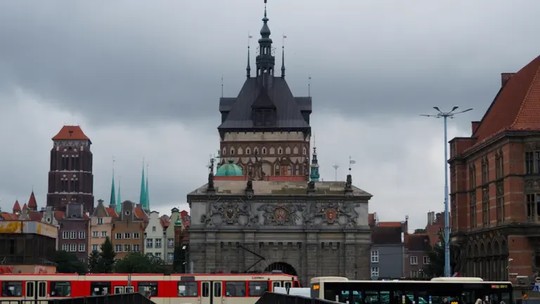 Czerwony tramwaj przejeżdża obok historycznej bramy w centrum Gdańska w pochmurny dzień.
